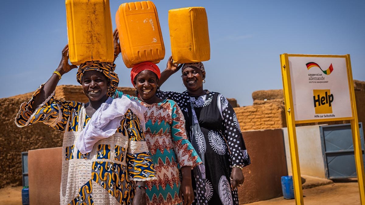 Drei Frauen stehen vor einem Help-Brunnen und tragen Wasserkanister auf den Köpfen