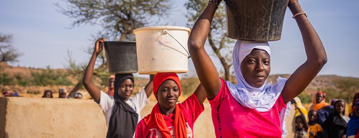 Drei malische junge Frauen tragen Wasser-Eimer auf dem Kopf