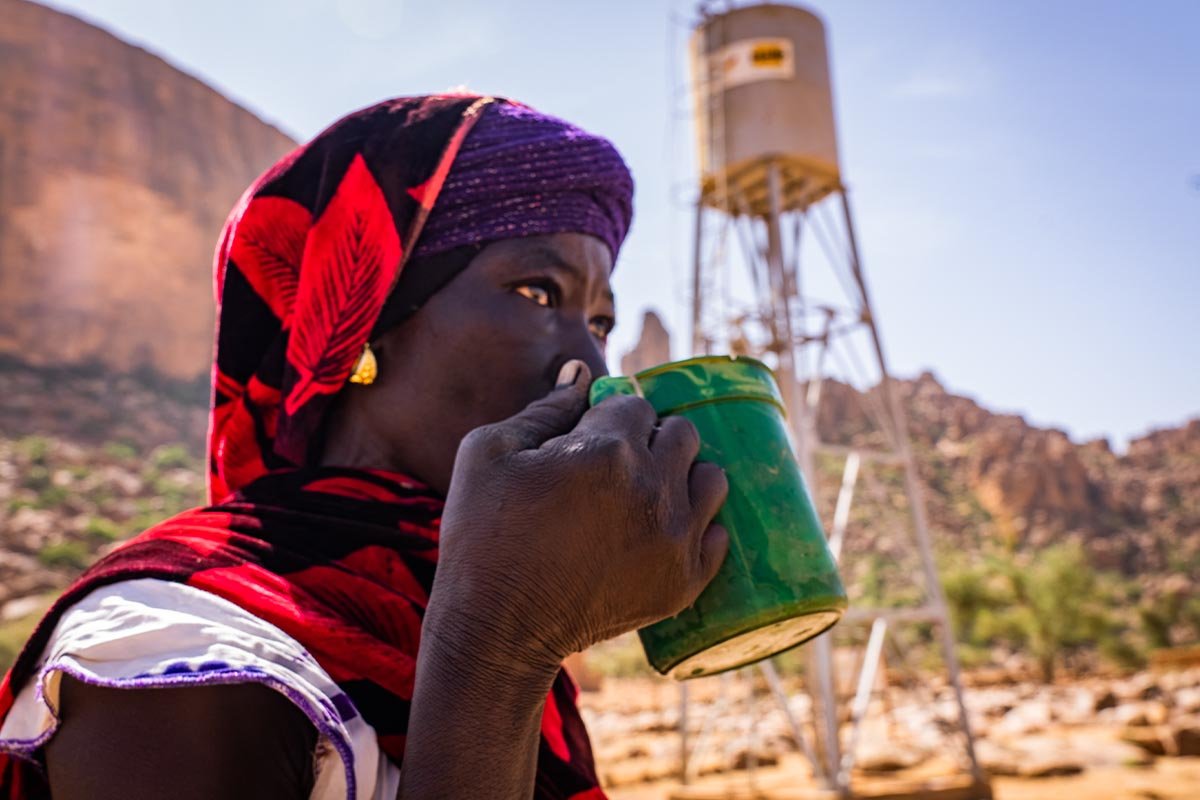 Eine Frau steht vor einem Help-Wasserturm und trinkt frisches Trinkwasser