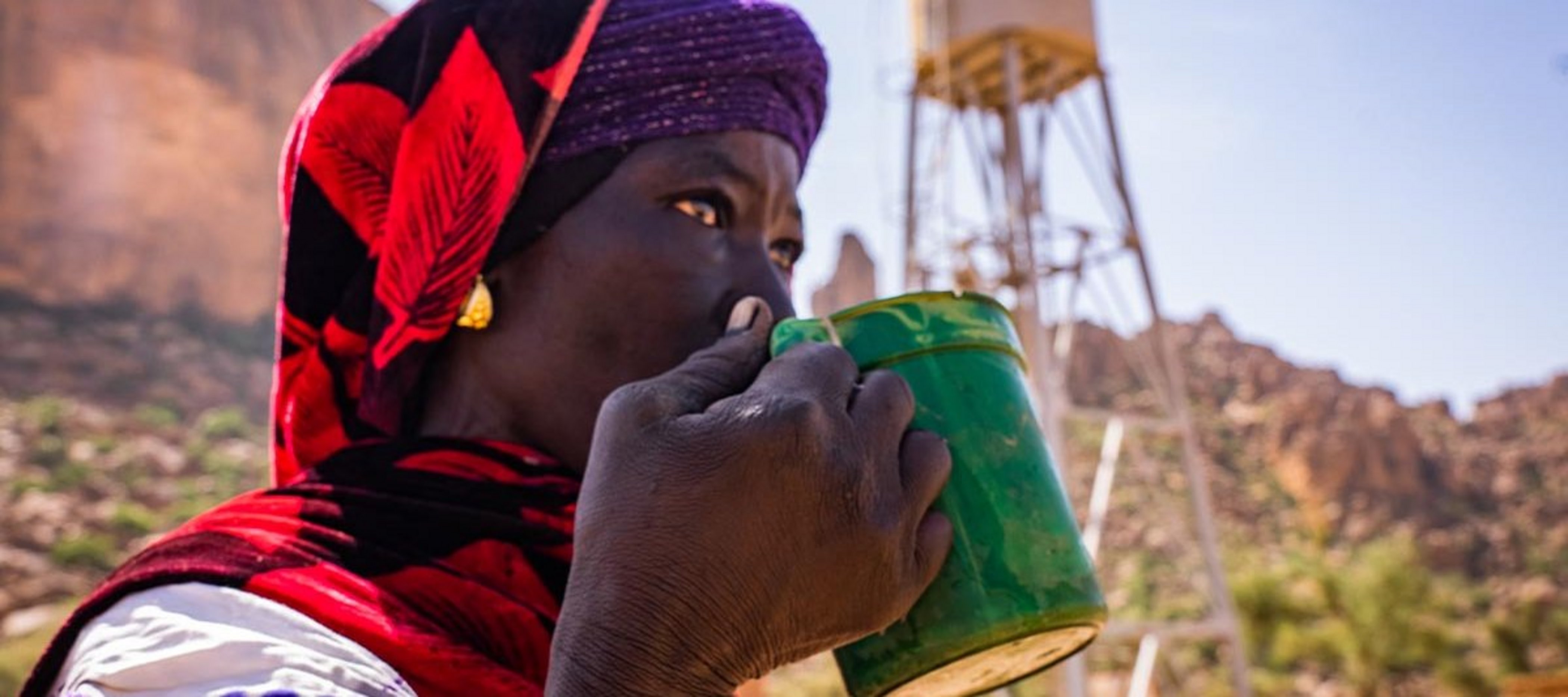 Eine Frau steht vor einem Help-Wasserturm und trinkt frisches Trinkwasser