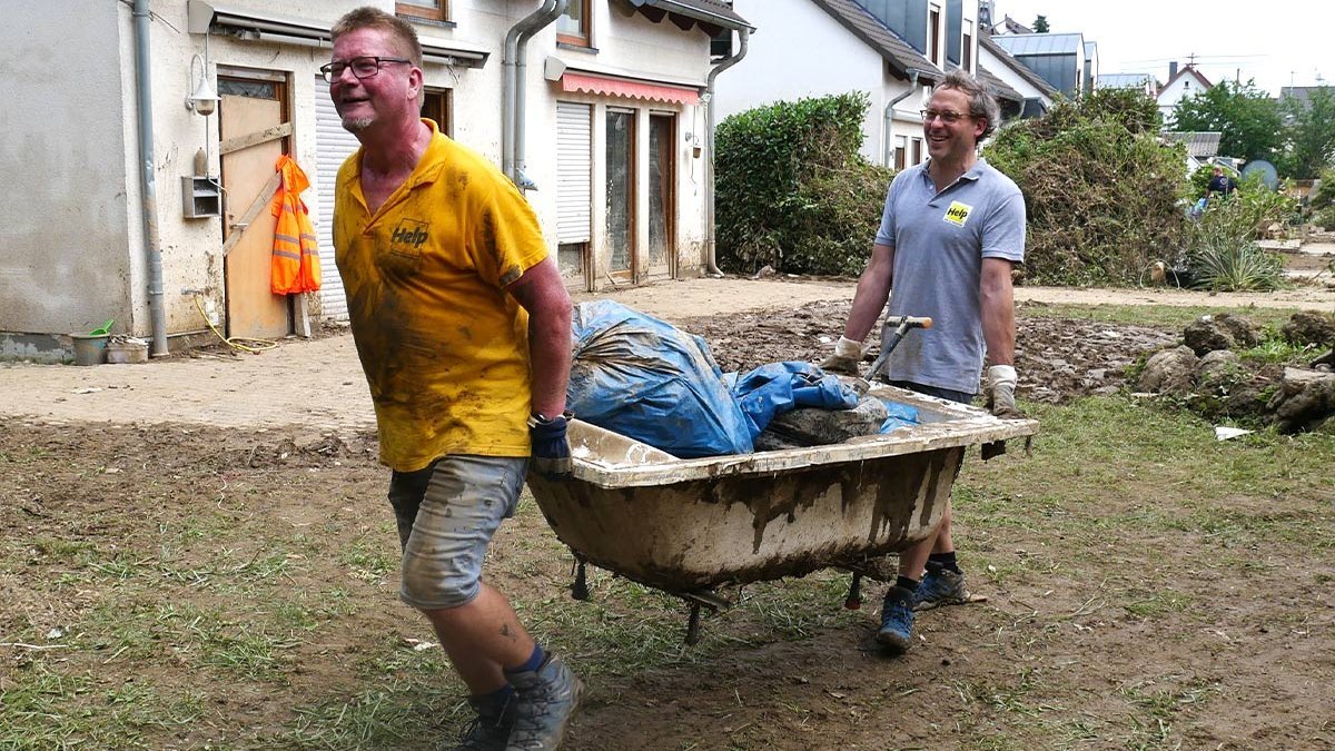 Zwei Help-Mitarbeiter tragen eine vom Hochwasser beschädigte Badewanne voller Schutt weg.