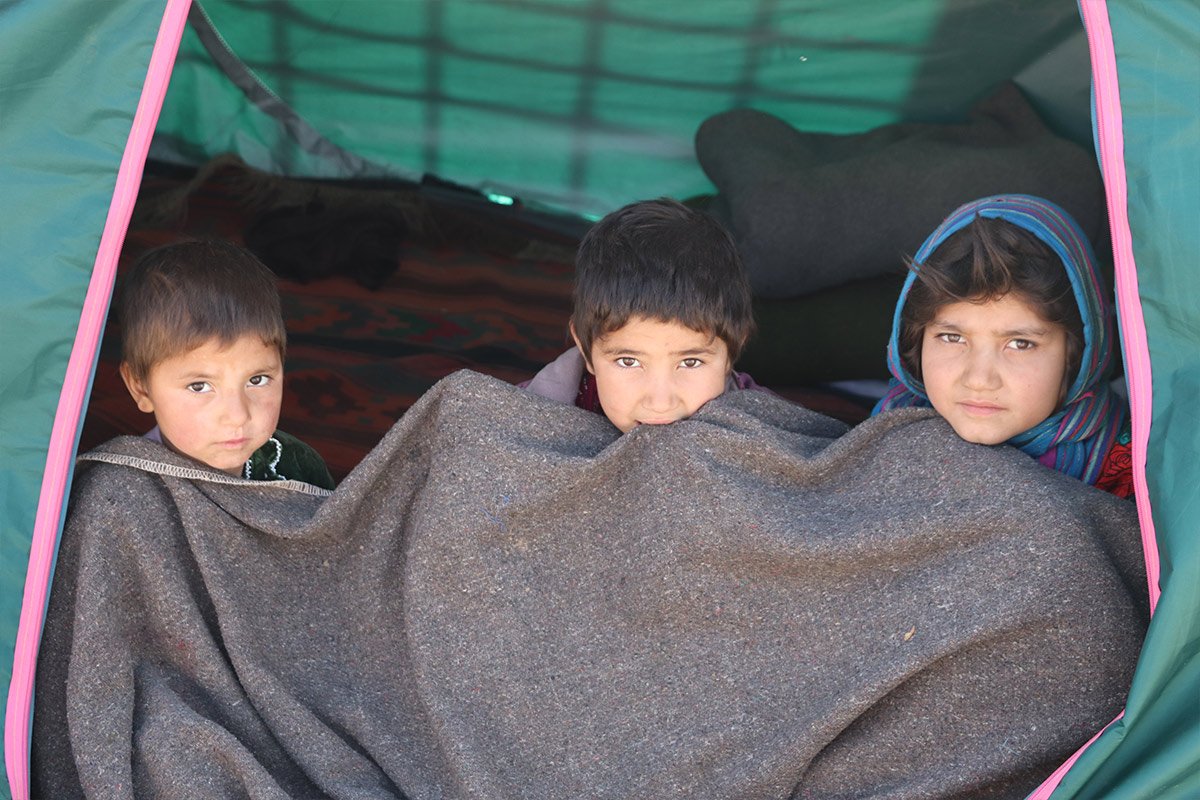 Drei Kinder aus Afghanistan sitzen in einem Flüchtlingszelt und versuchen, sich mit einer Decke zu wärmen.