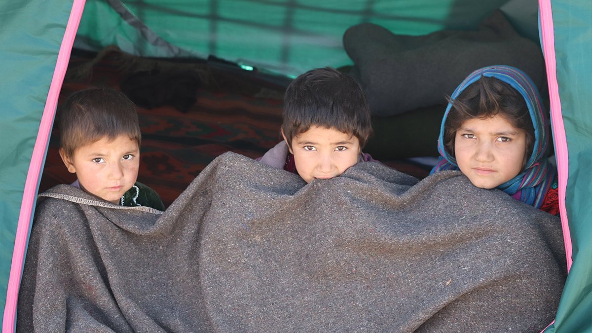 Drei Kinder aus Afghanistan sitzen in einem Flüchtlingszelt und versuchen, sich mit einer Decke zu wärmen.