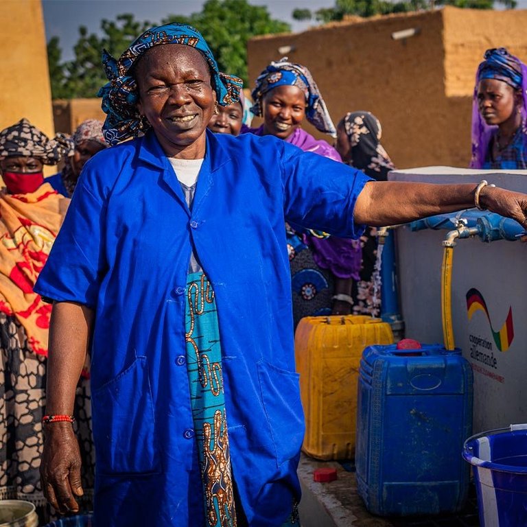Eine Frau in einem blauen Kleid steht vor einem Help-Brunnen und zapft Wasser ab
